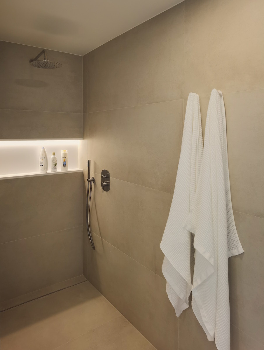 Ducha moderna grifería empotrada de acero cepillado, hornacina con iluminación indirecta acabada en marmol blanco y toallas blancas colgadas en pared beige.